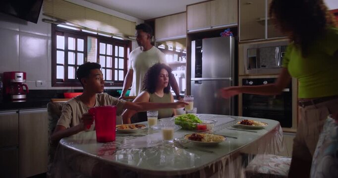 Family sitting down for lunch, mother, teenage son, and teenage daughter at the table with food and drinks, father standing in the background, enjoying a shared mealtime