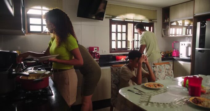 Teenage daughter serving spaghetti from the stove, mother assisting nearby, teenage son eating at the table, and father preparing food at the sink during family lunch