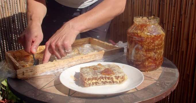 close view of hands spooning thick golden honey from jar onto honeycomb pieces on white plate, warm sunlit outdoor table, sticky texture golden highlights honey