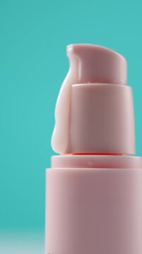 Woman's finger pressing a pink cosmetic pump dispenser bottle releasing face cream or lotion against a turquoise background