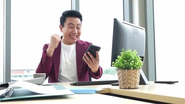 A happy man checks his smartphone at an office desk, reacts with excitement and celebrates a positive result while working beside a computer monitor and potted plant.
