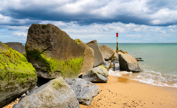 Massive weathered concrete sea defence blocks covered with vivid green algae at Horsey Gap on the Norfolk coast, England, with a red navigation marker, sandy beach and North Sea beneath a dramatic sum