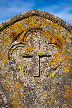 Close-up of a weathered stone grave marker with a carved Christian cross and circular motif, covered with vivid yellow-orange lichens in the historic churchyard at Rudston, East Yorkshire, England.