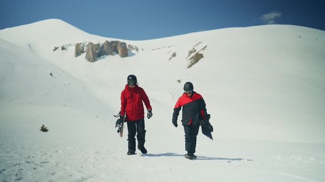 Two snowboarders trek uphill through a vast, snow-covered mountain landscape under a bright sky. Friends walk together in their winter sports gear, carrying their boards on a sunny day.