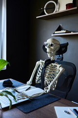 Skeleton wearing glasses and tie sitting at office desk with laptop, notebook and mouse