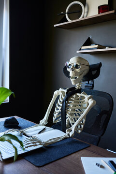 Skeleton wearing glasses and tie sitting at office desk with laptop, notebook and mouse