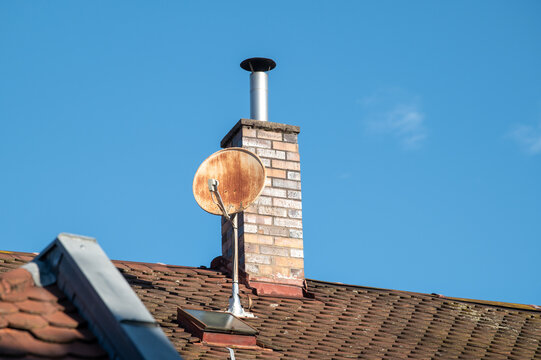 Untersicht verwitterte Satellitensch&uuml;ssel und gemauerter Schornstein aus Backsteinen mit Metallrohr auf Ziegeldach  in der b&uuml;rgerlichen Bergstadt in Bamberg vor blauem Himmel.	
