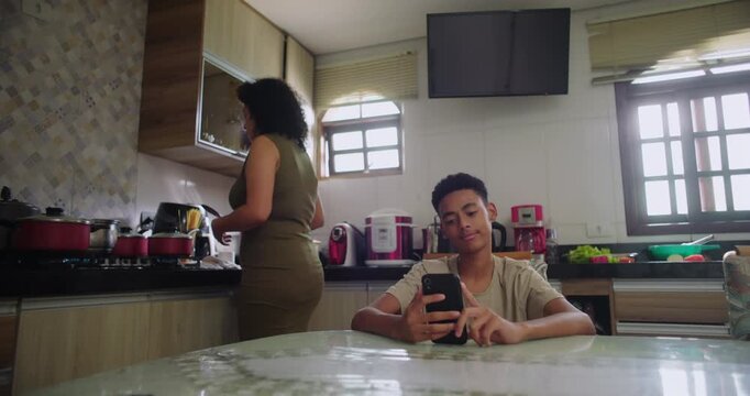 Teenage boy focused on smartphone while sitting at dining table as mother cooks at stove, family routine blending technology with daily meal preparation