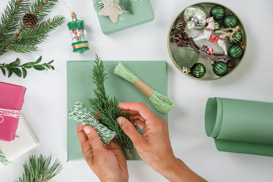Woman's hands holding a fir branch and decorative twine over a wrapped Christmas gift with assorted Christmas baubles and ornaments, wrapping paper and foliage on a white table