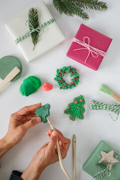 Personal perspective of a woman's hands shaping green air dry clay into a Christmas tree surrounded by wrapped Christmas gift boxes, twine, wooden tools and fir branches on a white table