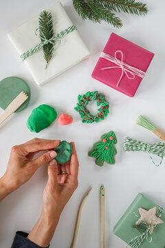 Personal perspective of a woman's hands shaping green air dry clay into a Christmas tree and wrapped Christmas gifts, twine, wooden tools and fir branches on a white table
