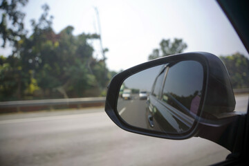 Side mirror reflects road and passing cars. Driver navigates highway, focused on journey. Blurry trees line the road behind. Image captures motion and travel in daylight. Ideal for themes of journey © Daniel Pawer
