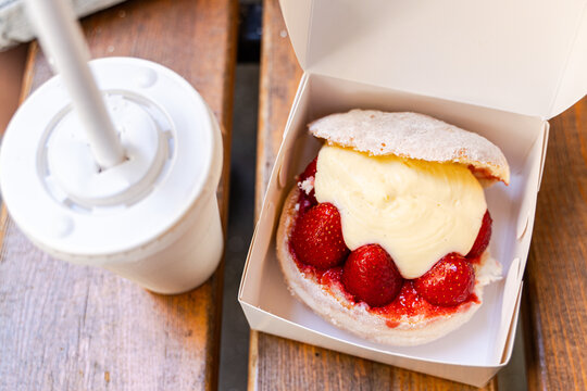 Close-up of a traditional Roman brioche bun filled with whipped cream and strawberries (Maritozzo) in a takeaway box on a wooden table with a takeaway drink