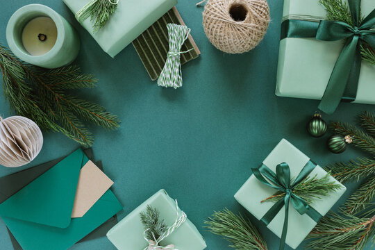 Overhead view of assorted wrapped Christmas gift boxes decorated with blank gift tags, evergreen sprigs, velvet ribbon, jute twine, Christmas baubles and ornaments on a green background