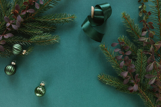 Overhead view of evergreen fir branches, barberry sprigs, a spool of ribbon and Christmas baubles on a green background