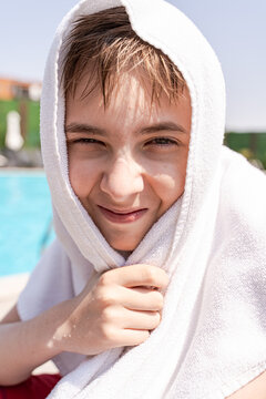 Close-up portrait of a smiling boy sitting by a swimming pool with a white towel on his head
