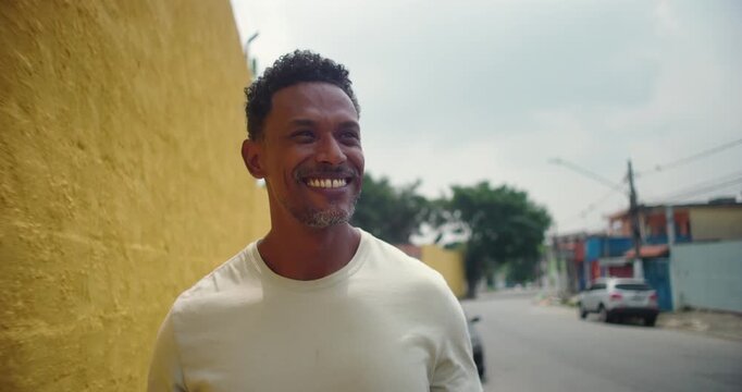 Smiling man of African descent walking along a vibrant yellow wall in an urban neighborhood, exuding positivity and confidence, with tropical plants in the background