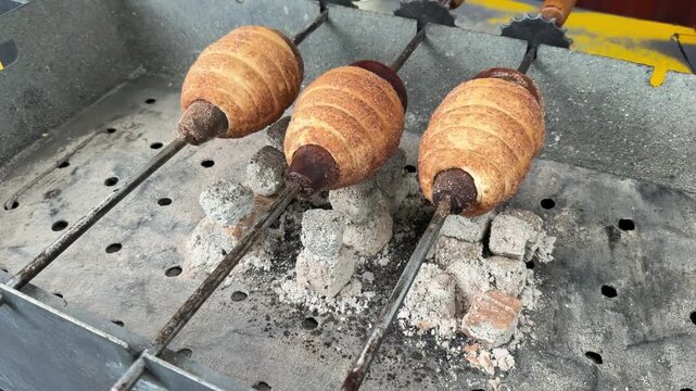 Trdelnik Three golden-brown pastries rotate on skewers above glowing embers in a metal grill, highlighting the cooking process in an outdoor environment