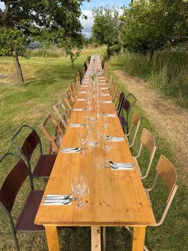 Long rustic wooden trestle table laid with wine glasses, cutlery, napkins and water carafes in a rural landscape on a summer day, UK