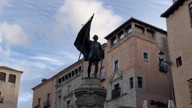 Low angle tracking shot of the bronze soldier statue holding a waving flag against historical medieval architecture in the sunny city center of Segovia, Spain.