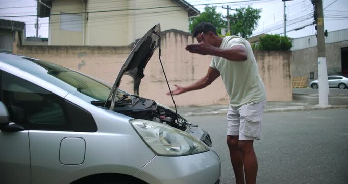 Man gesturing in frustration at broken car with hood open, expressing stress and struggle during mechanical failure in an urban street setting