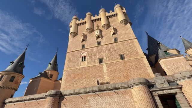 Low angle panning shot of the majestic Alcazar castle featuring ornate stone masonry and medieval turrets under a vibrant blue sky in Segovia, Spain.