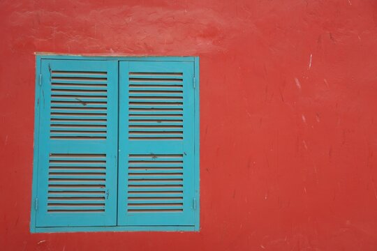 Vue rapproch&eacute;e de face d'une fen&ecirc;tre carr&eacute;e avec des volets en bois bleus ferm&eacute;s, au sein d'un mur peint en orange fonc&eacute;. Fa&ccedil;ade d'une maison tr&egrave;s color&eacute;e.
