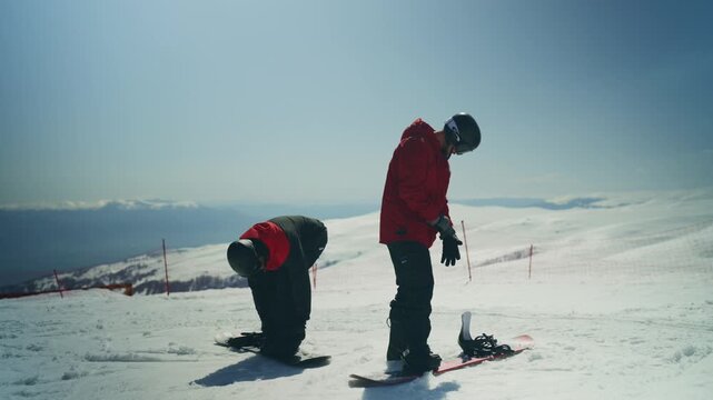 Two snowboarders are seen on a snowy mountain adjusting their boots and bindings. They are preparing for a thrilling day of winter sports under a clear sky.