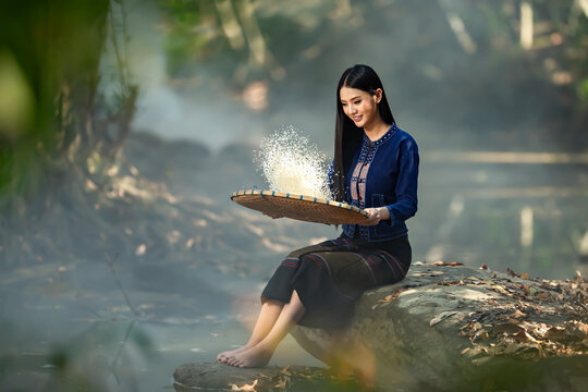 Smiling barefoot young woman in traditional Thai clothing sitting on a rock by a river winnowing rice (Fad Khao) using a bamboo tray (Kradong), Sakon Nakhon, Thailand