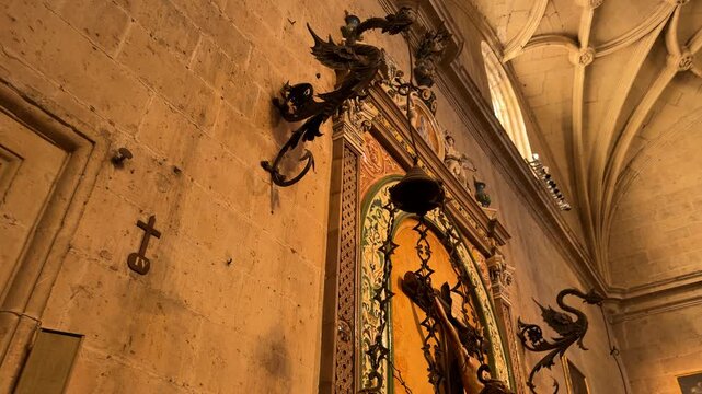 Low angle panning shot of the ornate crucifix and iron-wrought devotional hanging lamp inside the historic Segovia Cathedral, showcasing intricate medieval architecture in Segovia, Spain.
