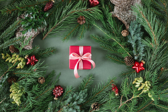 Overhead view of a wrapped Christmas gift surrounded by evergreen fir branches, thuja, pinecones and assorted festive red Christmas baubles and ornaments on a green background