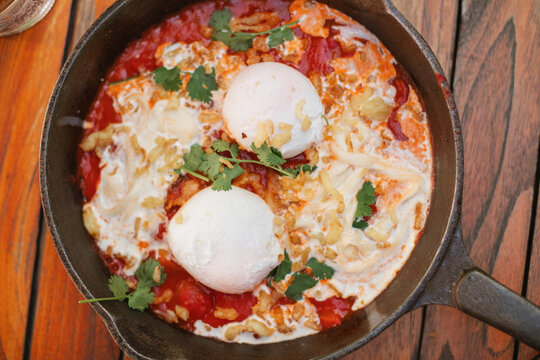 Close-up overhead view of a cast iron frying pan with tomato, poached eggs, stracciatella cheese and cilantro (shakshuka) on a wooden table