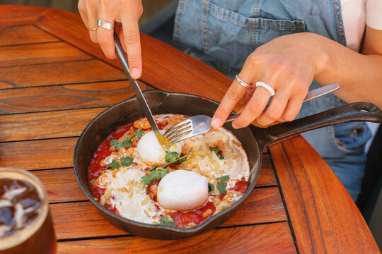 Close-up of a woman sitting at an outdoor cafe table eating poached eggs with tomato, stracciatella cheese and cilantro (shakshuka) with an iced espresso tonic coffee on a summer day
