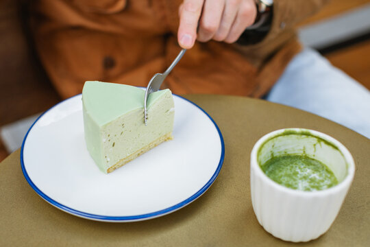 Close-up of a man sitting at an outdoor cafe table with a hot matcha latte drink cutting into a green matcha mousse cake with a fork