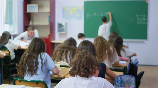 A group of young students sits at wooden desks in a bright classroom, while a classmate writes lesson notes on the large green chalkboard at the front of the room