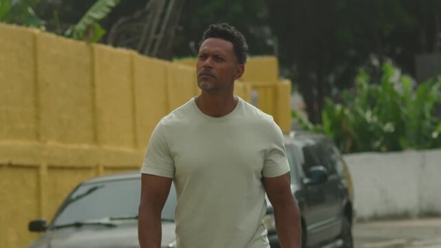 Man walking forward on urban street with pensive expression, focused gaze, strong posture, passing parked cars, yellow walls, and trees in background