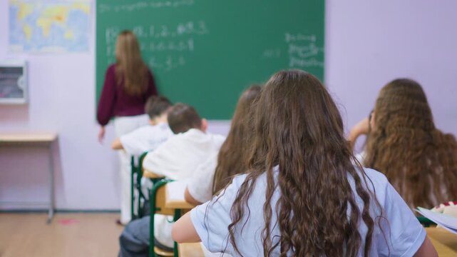 A group of young students sits at wooden desks in a bright classroom, while a classmate writes lesson notes on the large green chalkboard at the front of the room