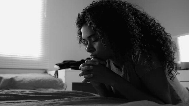 Teenage girl kneeling on bed in monochromatic scene, hands clasped, eyes closed, expressing deep prayer, spirituality, hope, faith, and emotional reflection