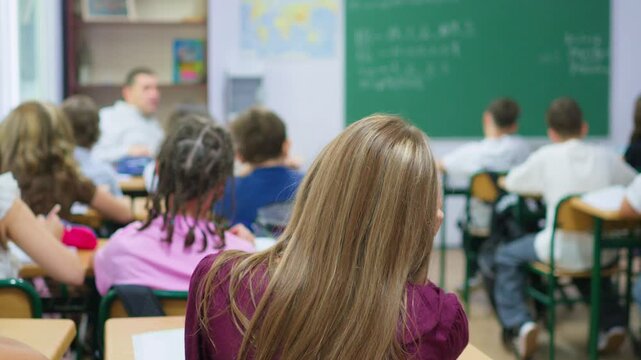 A group of young students sits at wooden desks in a bright classroom, while a classmate writes lesson notes on the large green chalkboard at the front of the room