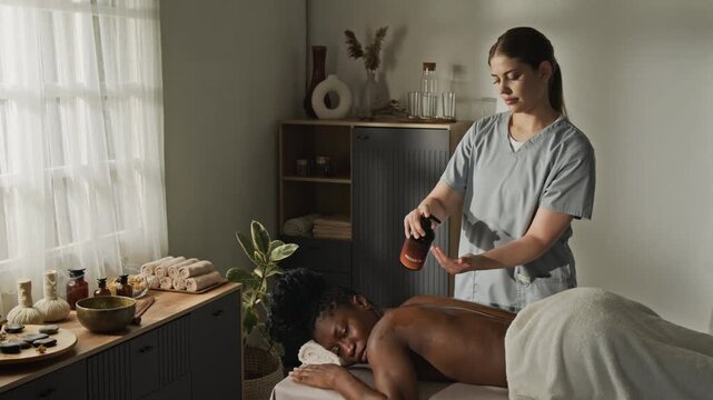 Caucasian massage therapist applying oil and massaging back of African American young adult woman. Ideal for wellness center marketing, spa clinic promo, or healthcare advertising