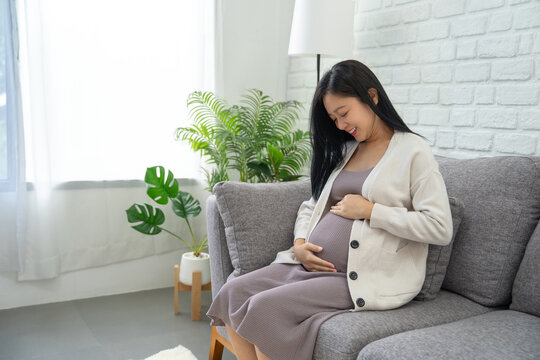 Happy pregnant Asian woman sitting on a sofa in the living room.