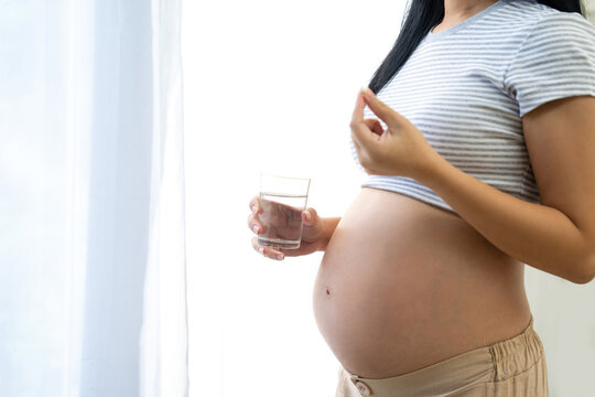 A pregnant woman is holding a bottle of water and some medicine, standing near a large window in her house.