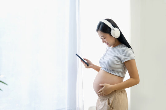 Pregnant Asian woman is listening to her favorite music on her smartphone while standing near a large window in her house.