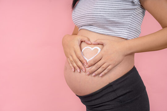 Pregnant young woman making heart with her hands on belly near window indoors