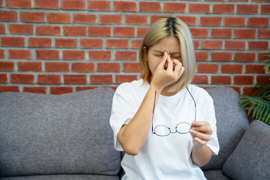 Asian woman is experiencing eye strain after working for long periods, holding her glasses while sitting on a sofa.