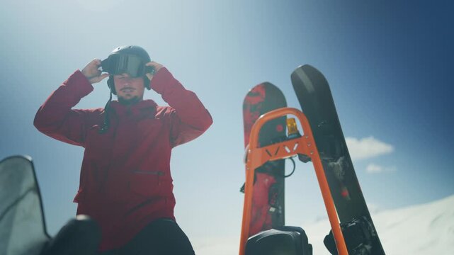 A man in winter attire adjusts his ski goggles, preparing for snowboarding. He stands beside a snowmobile with snowboards, ready for a mountain descent.