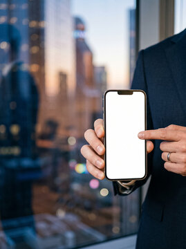 businessman holding smartphone with blank white screen near window with evening city view pointing finger at mockup for app presentation