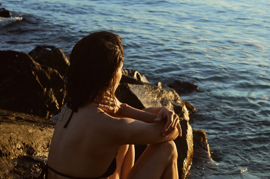 Woman sits on rocks by the water during sunset looking out at the sea