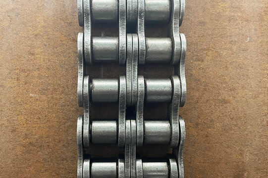 Heavy roller chain on brown metal surface background. Steel links arranged in straight rows. Industrial transmission component for machinery. Mechanical drive hardware in close detail.