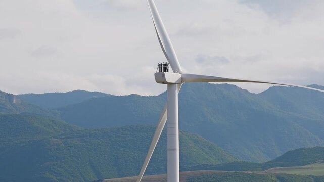 Technicians performing maintenance on a large wind turbine with rotating blades against a mountain landscape